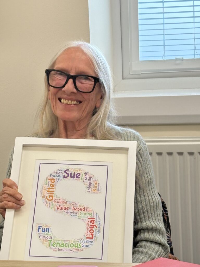 A woman with long grey hair and large dark glasses is smiling and holding a large framed print of a letter 'S'. The letter is made up of words which describe her character.