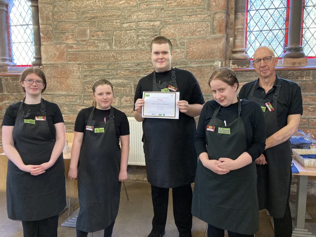 A young man is in the middle of the photo. He is holding a certificate with both hands, wearing a black uniform and an apron. He is smiling directly towards the camera. To the right is a young woman with brown hair in a ponytail, and to the right of her is a bald man, both of whom are also wearing the same black uniform and apron. To the left of the young man in the centre, there are two more young women, both of whom have brown hair in a ponytail. Everybody is smiling at the camera.